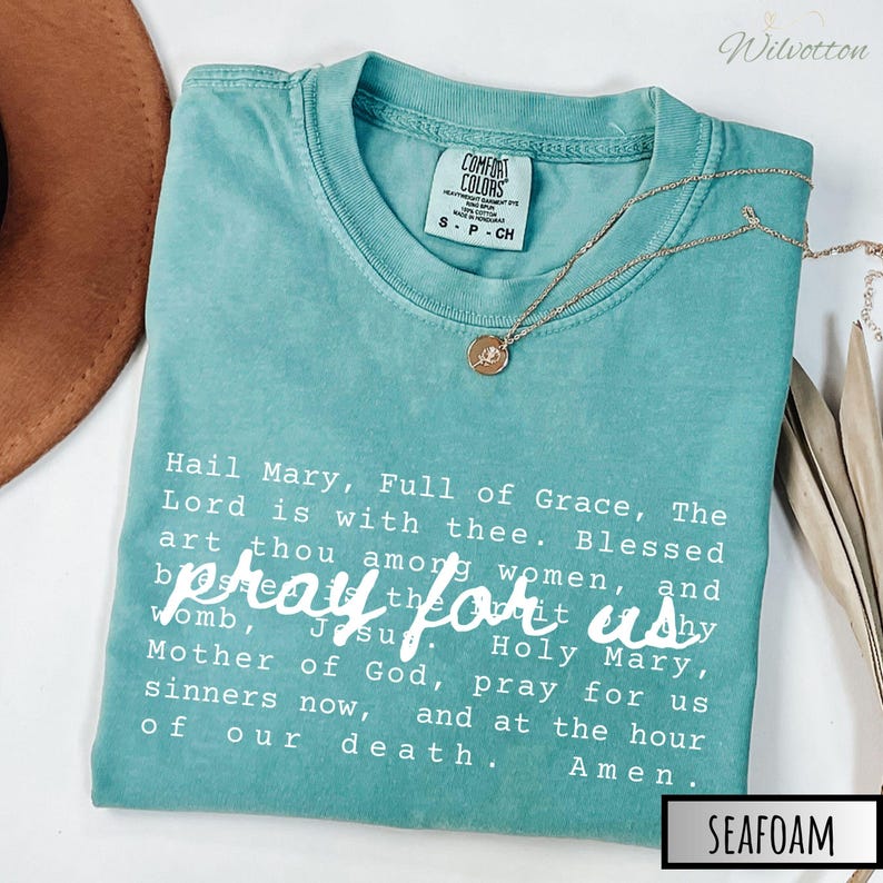 May include: Seafoam green Comfort Colors t-shirt with the text "Hail Mary, Full of Grace" and "pray for us" in white. A gold necklace with a pendant is draped on the shirt. The shirt is labeled "SEAFOAM".