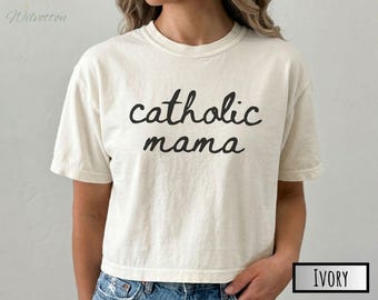 Puede incluir: Una camiseta de manga corta color marfil con el texto negro "catholic mama" impreso en letra cursiva en el pecho. La prenda tiene cuello redondo y un corte holgado, con la palabra "IVORY" mostrada en un pequeño recuadro negro abajo.