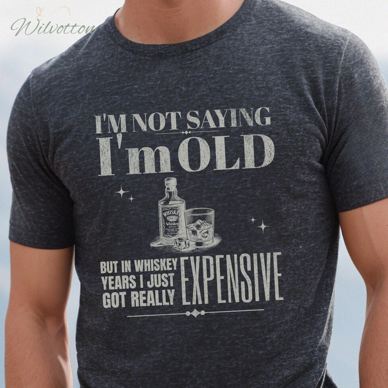 Puede incluir: Una camiseta gris oscura con un gr&aacute;fico blanco desgastado que dice "No estoy diciendo que soy viejo, pero en a&ntilde;os de whisky me he vuelto realmente caro". El gr&aacute;fico tambi&eacute;n incluye una botella de whisky y un vaso con cubitos de hielo.