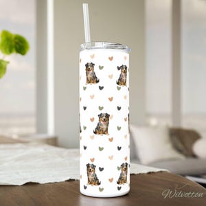 May include: A white tumbler with a lid and straw, featuring a repeating pattern of Australian Shepherd dogs and hearts in black, green, and tan. The tumbler is cylindrical and sits on a wooden surface.