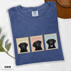 May include: Denim blue t-shirt with three postage stamp designs. Each stamp features a black Labrador portrait and the words "Forever Love." The shirt has a crew neck and a Comfort Colors tag. A brown hat is visible.