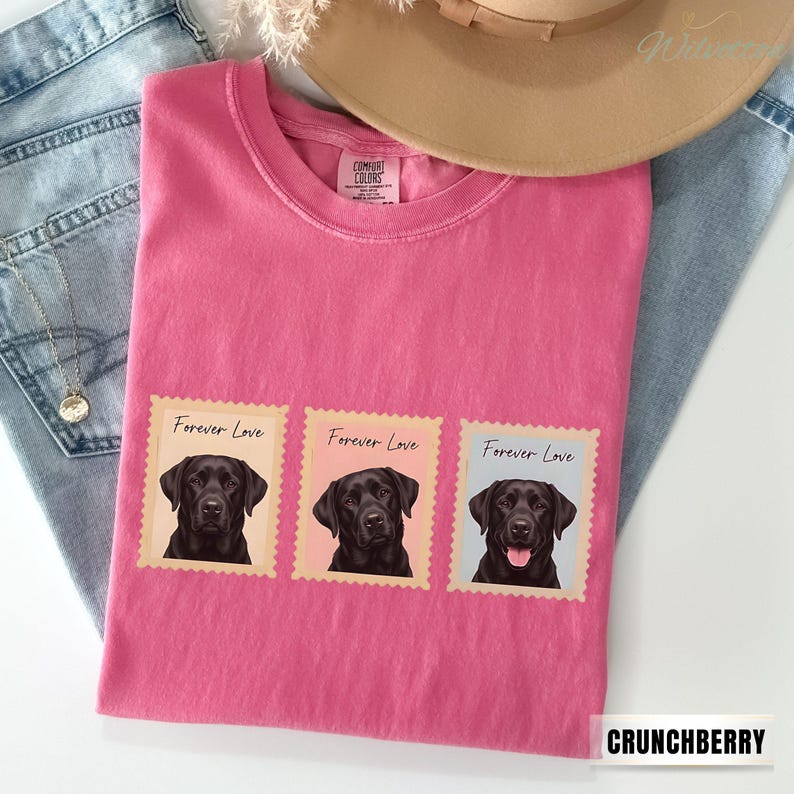 May include: A pink Comfort Colors t-shirt with three postage stamp designs. Each stamp features a black Labrador portrait and the words "Forever Love." The shirt is on a denim background with a hat.