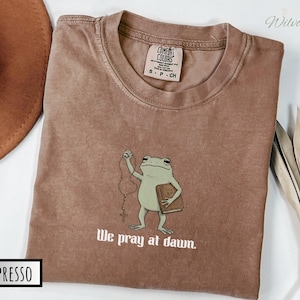 May include: An espresso-colored t-shirt featuring a graphic of a green frog holding a rosary and a book labeled Holy Bible. The text below the frog reads We pray at dawn. The shirt has a Comfort Colors brand label inside the collar.