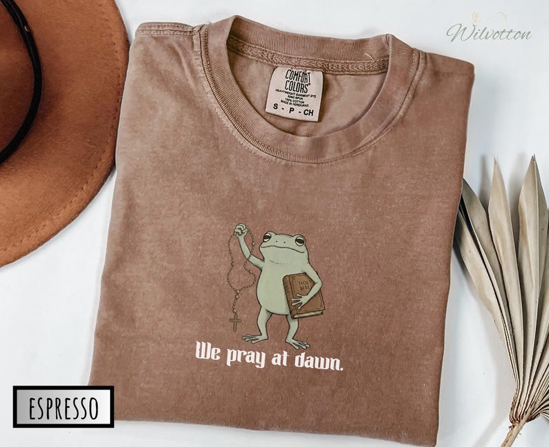 May include: An espresso-colored t-shirt featuring a graphic of a green frog holding a rosary and a book labeled Holy Bible. The text below the frog reads We pray at dawn. The shirt has a Comfort Colors brand label inside the collar.