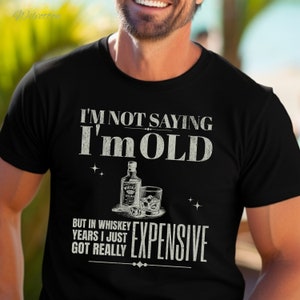 Puede incluir: Una camiseta negra con un gr&aacute;fico blanco que dice "No estoy diciendo que soy viejo, pero en a&ntilde;os de whisky me he vuelto realmente caro". El gr&aacute;fico tambi&eacute;n incluye una botella de whisky y un vaso de whisky con hielo.