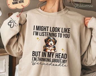 May include: A beige sweatshirt with the text "I might look like I'm listening to you, but in my head I'm thinking about my Bernedoodle". The sweatshirt features a cartoon Bernedoodle dog with brown, black, and white fur.