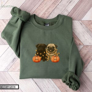 May include: Military green sweatshirt featuring a graphic of two pugs with string lights and pumpkins. The pumpkins are labeled "Lilly" and "Rex". The sweatshirt is a solid color with a crew neck and long sleeves.