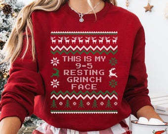 May include: A cardinal red sweatshirt with a festive design. The design includes reindeer, snowflakes, and Christmas trees, along with the text "THIS IS MY 9-5 RESTING GRINCH FACE". The sweatshirt is a comfortable, casual style.
