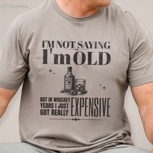 Puede incluir: Una camiseta gris con un dicho humor&iacute;stico sobre el envejecimiento. El texto dice "No estoy diciendo que soy viejo, pero en a&ntilde;os de whisky me he vuelto realmente caro". La camiseta presenta una ilustraci&oacute;n estilizada de una botella de whisky y un vaso con cubitos de hielo.
