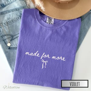 Puede incluir: Una camiseta violeta con las palabras "made for more" en escritura blanca y un lazo gr&aacute;fico. La camiseta est&aacute; doblada y colocada encima de unos vaqueros azules y un sombrero marr&oacute;n. La camiseta est&aacute; etiquetada como "Violeta".
