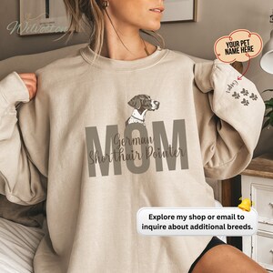 May include: Beige sweatshirt with the word "MOM" in large gray letters, and "German Shorthair Pointer" underneath. A graphic of a German Shorthair Pointer is above the text. The sleeve has a pet's name and paw prints.