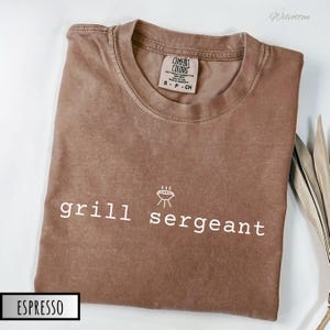 May include: A brown t-shirt with the text "grill sergeant" and a small grill graphic in white. The shirt is made of a soft, comfortable material and has a distressed look.