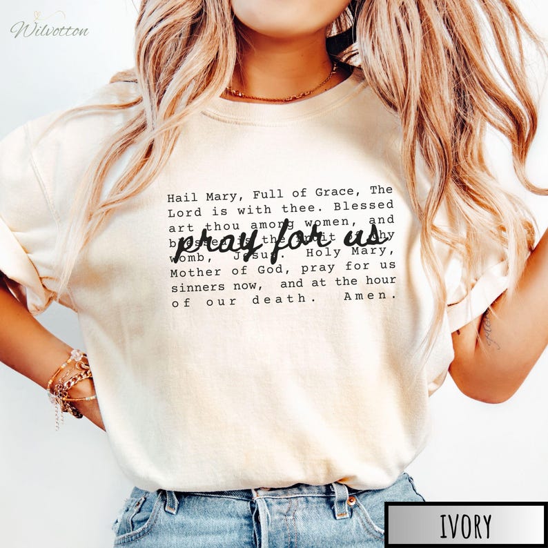 May include: Ivory-colored crew neck t-shirt with the text "Hail Mary, Full of Grace, The Lord is with thee...pray for us...Amen" in black. The shirt is a neutral color and features a classic, religious quote.
