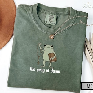 May include: A moss green crew neck t-shirt featuring a graphic of a light green frog holding a brown book labeled Holy Bible and a rosary. The text below the frog reads We pray at dawn. The shirt has a Comfort Colors brand label inside the collar.