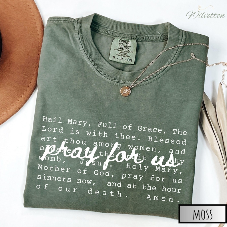 May include: A moss green Comfort Colors t-shirt with the text "Hail Mary, Full of Grace" and "pray for us" in white. A gold necklace with a pendant rests on the shirt. The shirt is a size small.