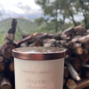 May include: A white candle with a copper-colored lid. The candle has the words "CRAWFORD CANDLES" and "Mountain Monsoon" printed on it. The candle is 10 oz. and is scented soy. The background is blurred, showing a natural outdoor setting.