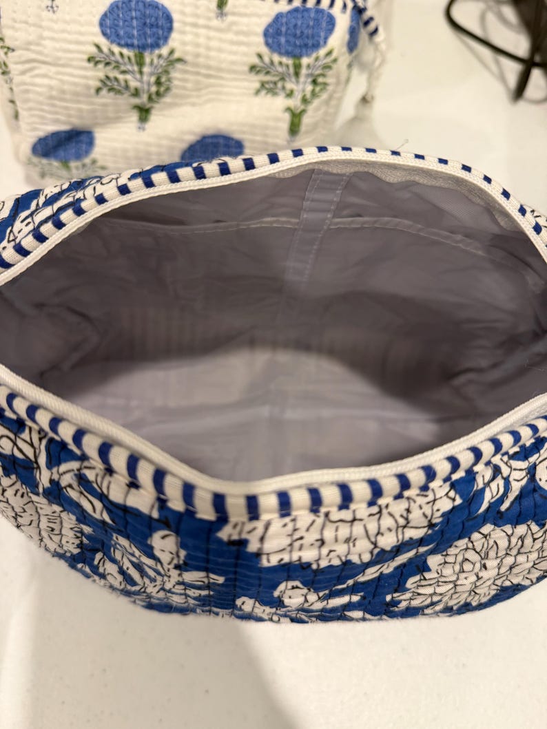 May include: A blue and white floral patterned cosmetic bag with a white zipper and a gray interior. The bag is open, revealing the inside. Another bag with a similar floral pattern is visible in the background.