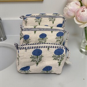 May include: Three cream-colored cosmetic bags stacked on a white surface. Each bag features a blue floral pattern and a navy blue striped trim. The bags have a zipper closure and a small tassel.