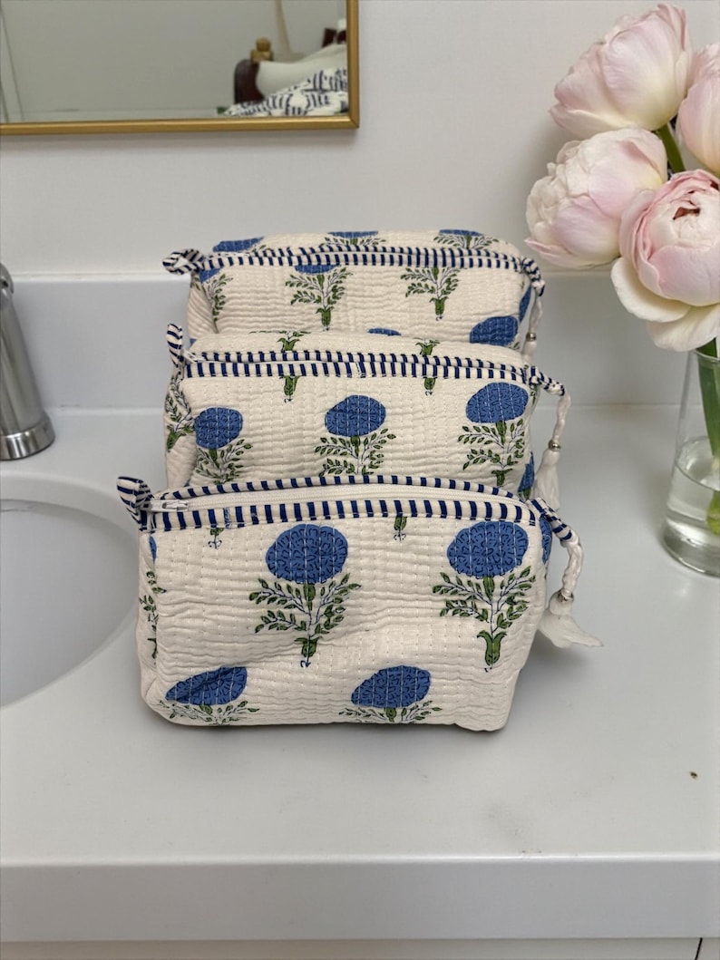 May include: Three cream-colored cosmetic bags stacked on a white surface. Each bag features a blue floral pattern and a navy blue striped trim. The bags have a zipper closure and a small tassel.