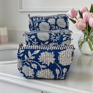 May include: Three blue cosmetic bags with white floral patterns are stacked on a white surface. The bags have a quilted texture and a striped border. A vase of pink tulips is in the background.