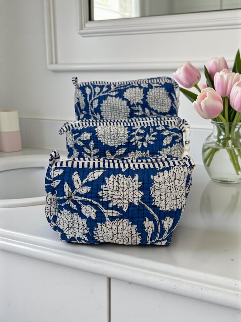 May include: Three blue cosmetic bags with white floral patterns are stacked on a white surface. The bags have a quilted texture and a striped border. A vase of pink tulips is in the background.