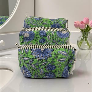 May include: Three quilted cosmetic bags stacked on a white surface. The bags feature a green background with blue floral patterns. Each bag has a black and white striped trim and a white tassel on the zipper.
