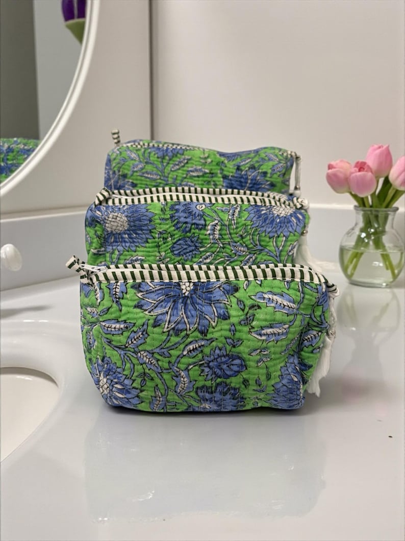 May include: Three quilted cosmetic bags stacked on a white surface. The bags feature a green background with blue floral patterns. Each bag has a black and white striped trim and a white tassel on the zipper.