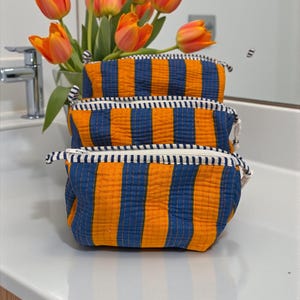 May include: Three orange and blue striped cosmetic bags with white zippers are stacked on a white surface. A bouquet of orange tulips is in the background. The bags have a quilted texture and are made of fabric.