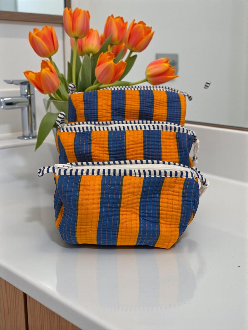 May include: Three orange and blue striped cosmetic bags with white zippers are stacked on a white surface. A bouquet of orange tulips is in the background. The bags have a quilted texture and are made of fabric.
