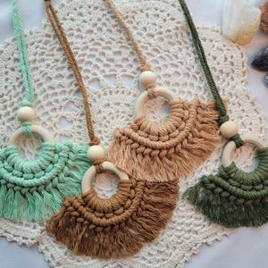May include: Four macrame car air fresheners in shades of green and brown. Each has a wooden ring and bead, with a fringed fan design. The air fresheners are displayed on a white crocheted doily, with crystals in the background.