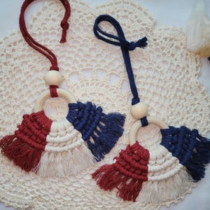 May include: Two macrame car air fresheners with red, white, and blue tassels. Each has a wooden ring and bead, and a matching cord for hanging. The air fresheners are displayed on a cream-colored, crocheted doily.