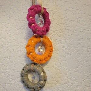 May include: A macrame wall hanging featuring three circular woven elements in shades of pink, orange, and green. Each ring has a crystal pendant hanging from the center. The piece is suspended by a natural fiber cord and a wooden bead.