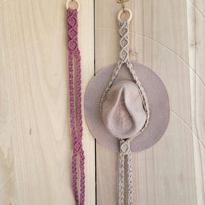 May include: Two macrame hat hangers with brass hooks are displayed against a light wood background. One hanger is a dusty rose color, and the other is a neutral beige. A wide-brimmed hat hangs from the beige hanger.