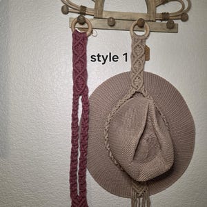 May include: A macrame wall hanging with a wooden sunburst design and a wooden bar with pegs. A mauve macrame strap hangs from a wooden ring, and a beige hat hangs from another ring. The text "style 1" is visible.