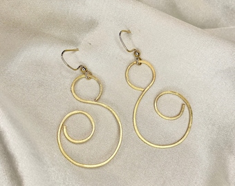 May include: A pair of gold-colored swirl earrings. Each earring features a small circle at the top, connected to a larger, elongated swirl design. The earrings are displayed on a cream-colored, textured fabric.