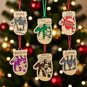 Op de afbeelding: Houten kerstversieringen in de vorm van wanten, elk met een uniek ontwerp en boodschap. De ornamenten tonen uitgesneden afbeeldingen van een hert, sneeuwman, kerstman en peperkoekmannetje. Boodschappen zijn "Hope", "Let it Snow", "Happy Holidays", "Believe", "Merry Christmas" en "Love".