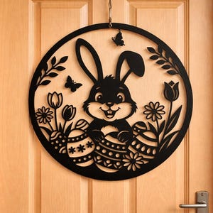 Op de afbeelding: Zwarte cirkelvormige paasdecoratie met een lachend konijn dat paaseieren vasthoudt. Het ontwerp omvat bloemen, vlinders en bladeren uit één stuk materiaal gesneden. De decoratie hangt aan een houten deur.