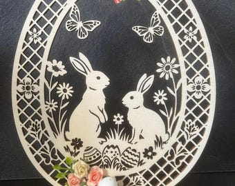 Puede incluir: Decoración de huevo de Pascua de madera con dos conejos, mariposas y flores. El marco en forma de huevo tiene un diseño de celosía. Un lazo a cuadros rojo y blanco y flores artificiales adornan la parte superior. Un huevo de Pascua con una cinta a cuadros roja y blanca está en el frente.