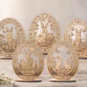 Może przedstawiać: Pięć drewnianych dekoracji wielkanocnych w kształcie jajek z wycinanymi laserowo wzorami. Każde jajko przedstawia królika, wzory kwiatowe i dekoracyjną obwódkę. Niektóre jajka zawierają napis "Happy Easter". Jajka stoją na drewnianych podstawach.