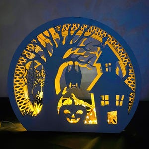 May include: A Halloween-themed, layered wooden decoration. The design features a spooky house, a tree with a bat, a jack-o'-lantern, and a spider web. The decoration is illuminated with warm yellow lights, creating a festive glow.
