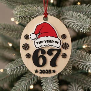 May include: A wooden Christmas ornament with a red Santa hat and the text "THE YEAR OF 67" in black. The year "2025" is below the number 67. The ornament is decorated with black snowflakes and hangs from a red string.