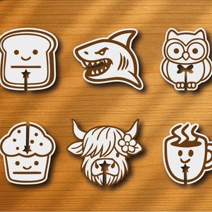 Op de afbeelding: Een verzameling van zes witte en bruine stickers met cartoon illustraties. De ontwerpen omvatten een sneetje brood, een haai, een uil, een cupcake, een Schotse hooglander en een koffiemok. Geplaatst op een houten oppervlak.