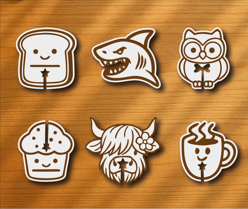 Op de afbeelding: Een verzameling van zes witte en bruine stickers met cartoon illustraties. De ontwerpen omvatten een sneetje brood, een haai, een uil, een cupcake, een Schotse hooglander en een koffiemok. Geplaatst op een houten oppervlak.