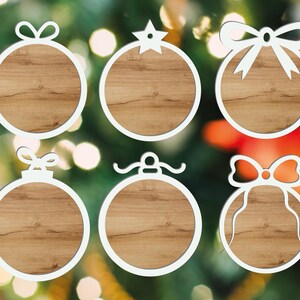 Puede incluir: Seis adornos navide&ntilde;os con marco blanco y centros con aspecto de madera. Cada adorno tiene una parte superior decorativa diferente, incluyendo lazos y una estrella. Los adornos se colocan sobre un fondo borroso de luces y vegetaci&oacute;n navide&ntilde;as.