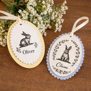 May include: Two Easter egg-shaped gift tags with scalloped edges. One is yellow, the other blue. Each tag features a black and white illustration of a rabbit and the names "Oliver" and "Claire". Cream-colored ribbon loops are attached.