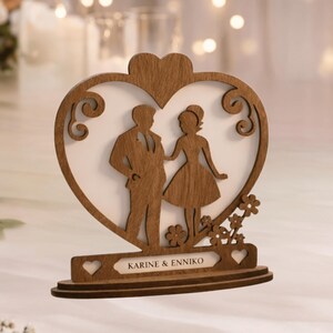 May include: A wooden wedding cake topper featuring a bride and groom silhouette inside a heart-shaped frame. The base includes the names "KARINE & ENNIKO" and decorative floral accents. The topper is made of light brown wood.