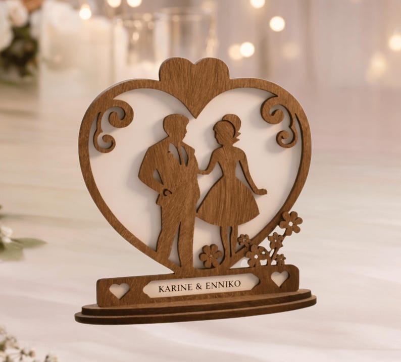 May include: A wooden wedding cake topper featuring a bride and groom silhouette inside a heart-shaped frame. The base includes the names "KARINE & ENNIKO" and decorative floral accents. The topper is made of light brown wood.
