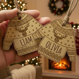 Puede incluir: Dos adornos navideños de madera con forma de suéter, con los nombres "LIAM" y "OLIVIA" grabados. Cada adorno presenta un diseño de reno y detalles de copos de nieve. Colgados de una cuerda, listos para colgar en el árbol de Navidad.