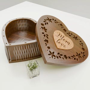Puede incluir: Una caja de madera en forma de corazón con tapa abierta. La caja es marrón con una inserción en forma de corazón de madera más clara en la tapa que dice "Mom love". La tapa tiene un diseño floral tallado en la madera. La caja está abierta, revelando el interior.