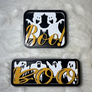 Peut inclure: Deux décorations rectangulaires noires d'Halloween avec des découpes de fantômes blancs et le mot "Boo!" en script doré. Une décoration a deux fantômes, l'autre en a quatre. Les décorations sont sur une surface blanche et moelleuse.